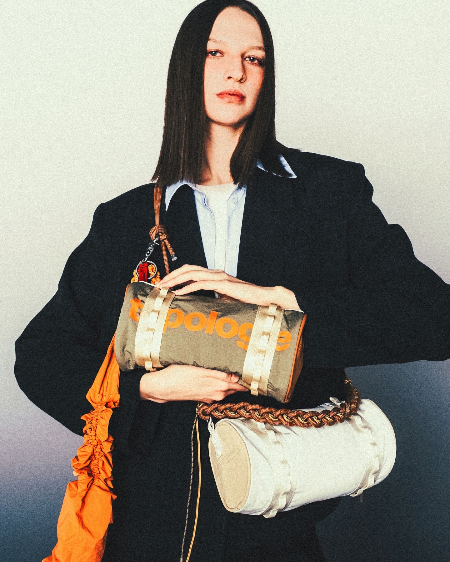 Fashion model in oversized black blazer holding two cylindrical designer bags — khaki bag with orange logo and cream bag, studio portrait