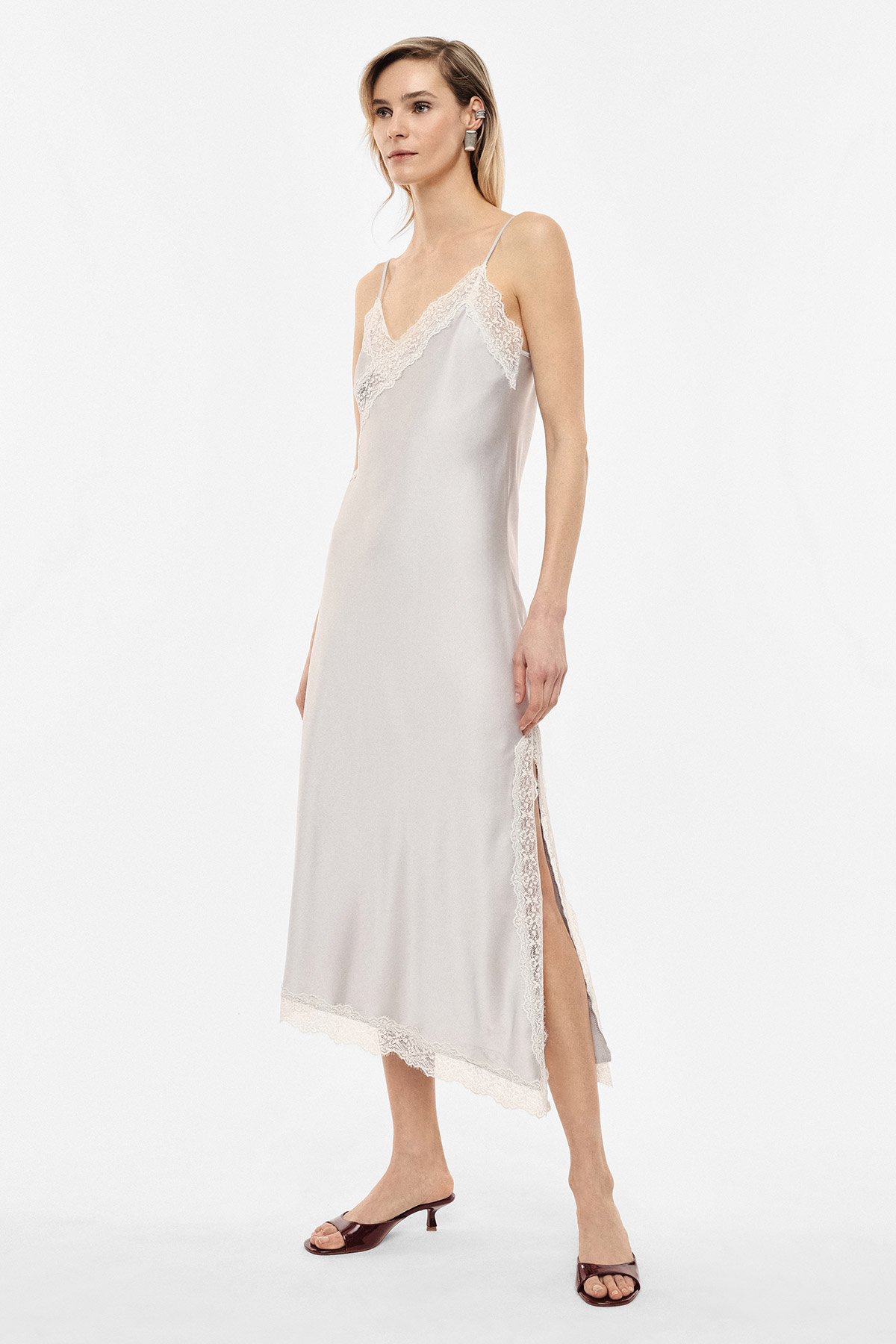 Model wearing a pale silk slip dress with lace trim, spaghetti straps and side slit on a white background
