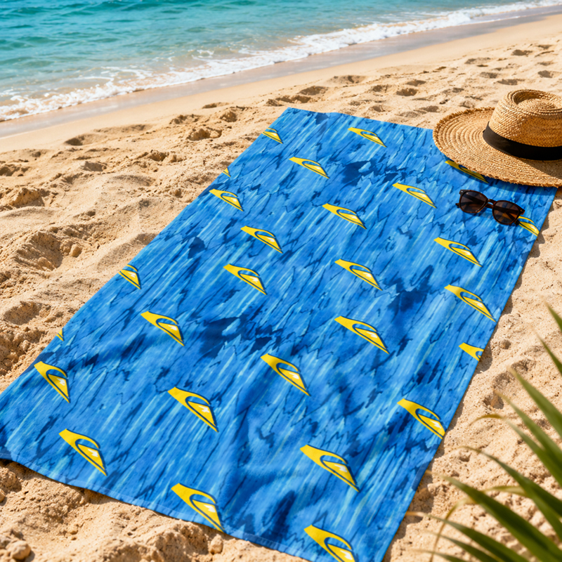 Blue patterned beach towel with yellow sail motifs laid on sandy shore beside straw hat and sunglasses, turquoise sea in background