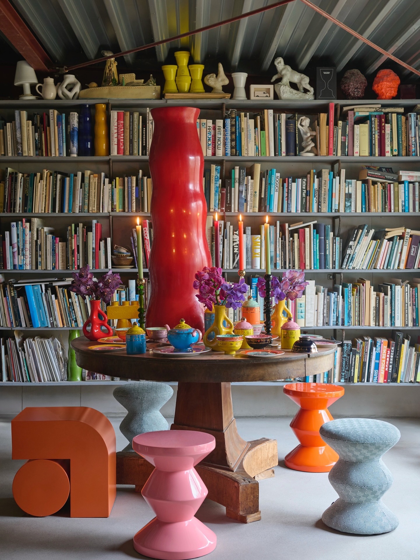 Eclectic reading room with floor-to-ceiling bookshelves, round wooden table topped by a tall red vase, lit candles, colourful ceramics and stools