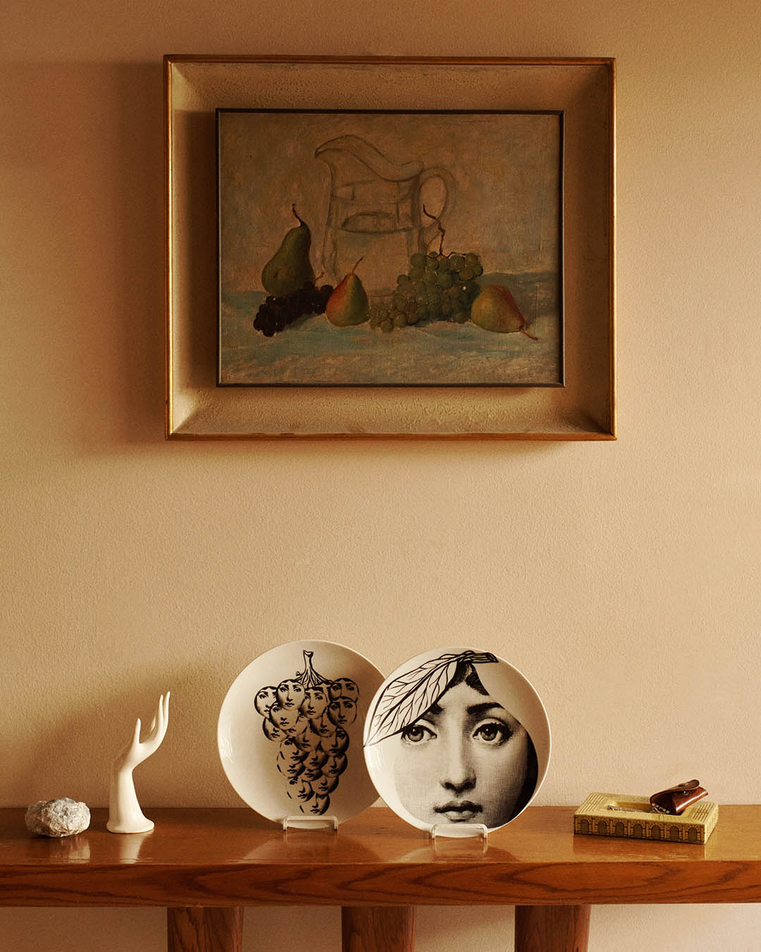 Framed still-life painting above a wooden shelf displaying two decorative porcelain plates (one with a face motif), ceramic hand, rock and book.