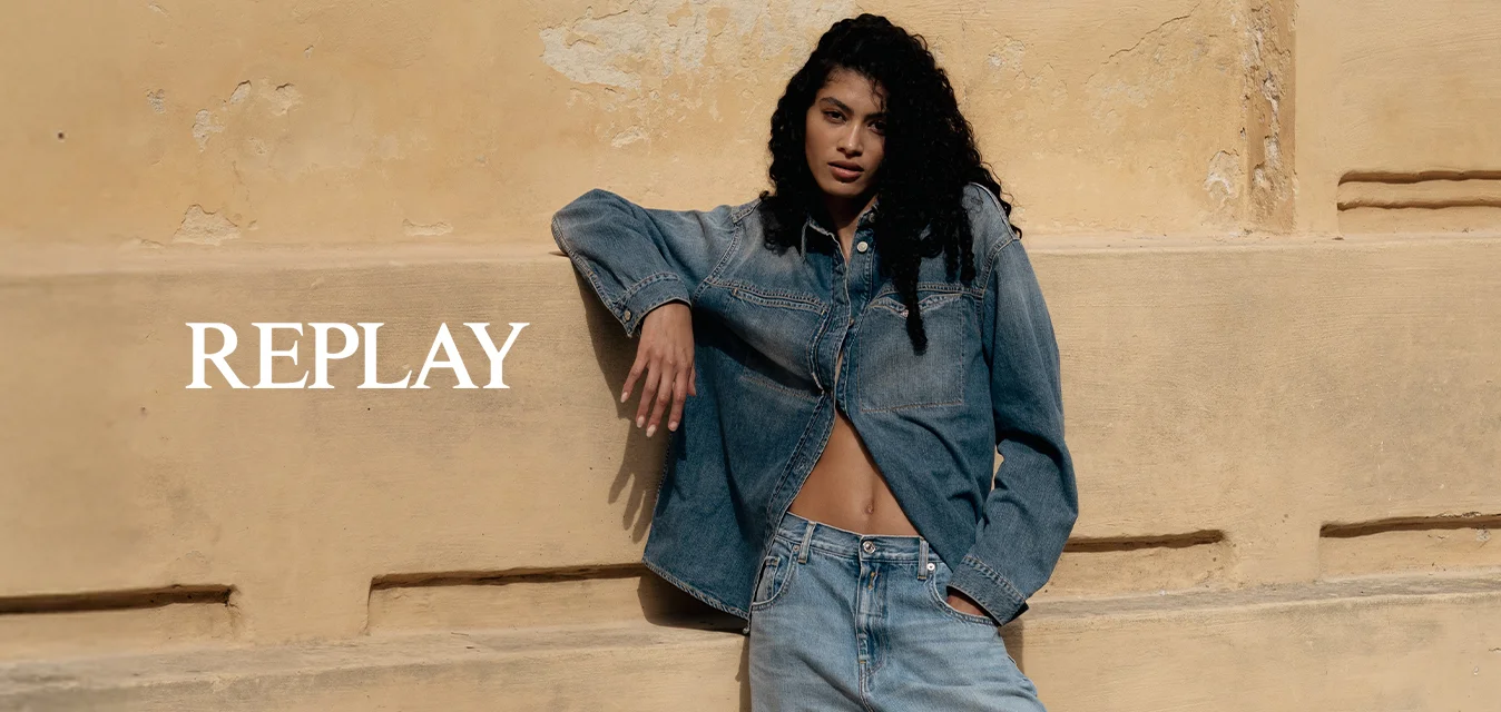Donna con capelli lunghi e ricci appoggiata a un muro beige testurizzato, indossa una giacca di jeans, jeans larghi e sneakers beige all'aperto.