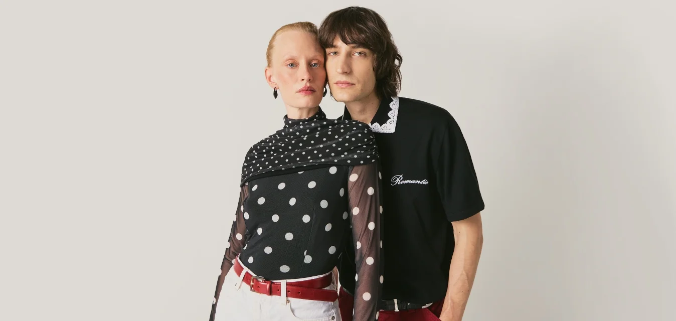 Due giovani adulti si tengono vicini; uno indossa una maglia trasparente a pois con pantaloncini bianchi, l'altro una polo nera e pantaloni rossi su uno sfondo semplice.