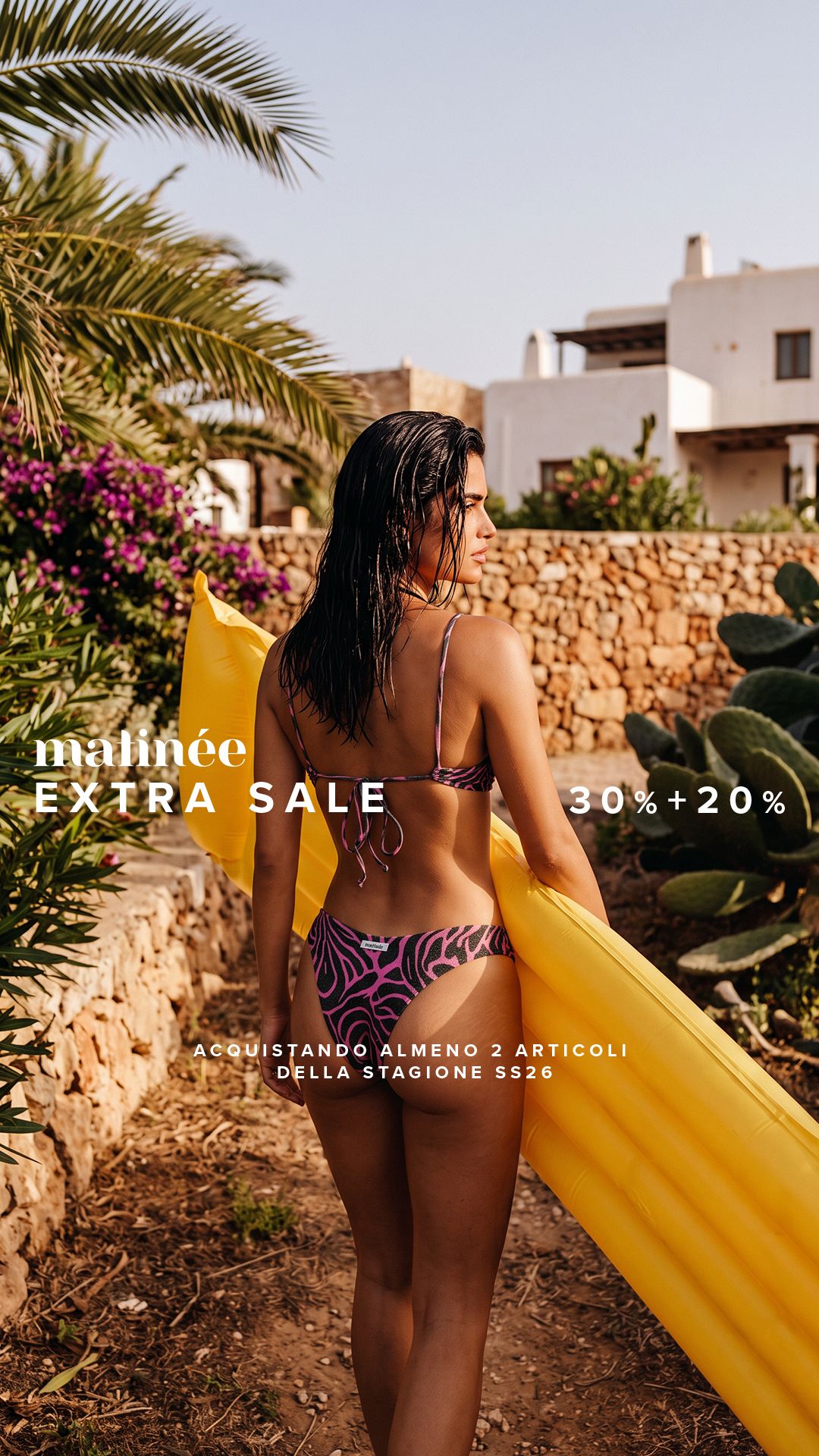 Woman in pink patterned bikini carrying a yellow inflatable mattress along a sunlit stone path past cacti and white holiday villas
