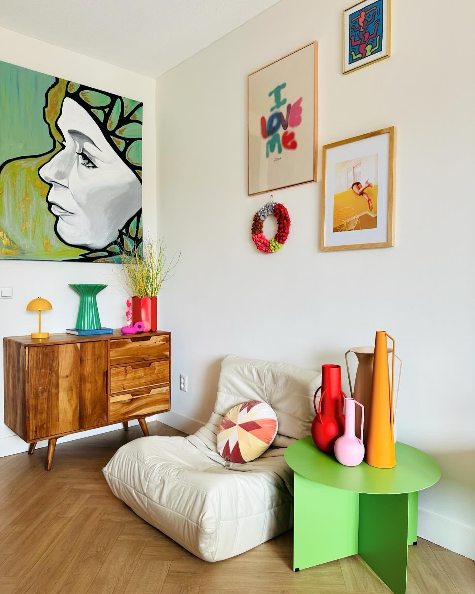 Colourful living room corner with low white lounge chair, wooden sideboard and green table with bright vases and framed art