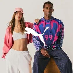 Una donna con pantaloni bianchi, crop top e giacca rosa sta accanto a un uomo seduto su uno sgabello che indossa una felpa con cappuccio colorata e pantaloni scuri.