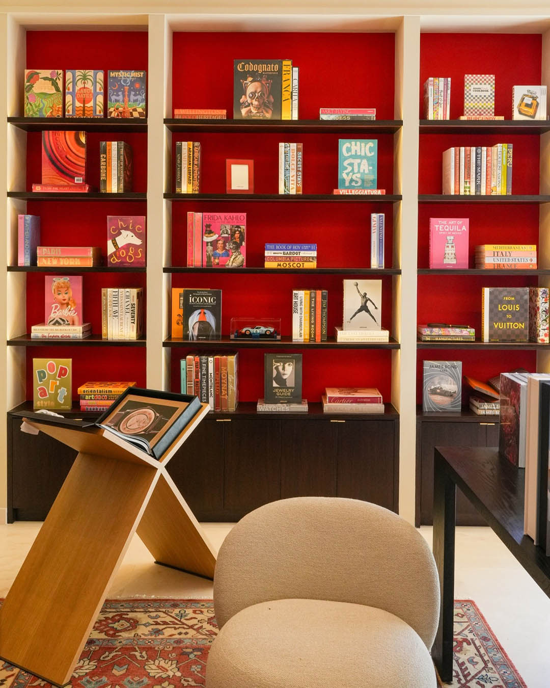 Cosy reading nook with red-back bookshelf filled with colourful books, modern wooden bookstand, rounded beige armchair