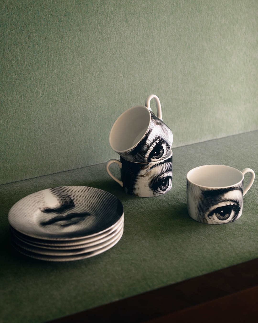 Stacked porcelain plates and mugs printed with vintage stylised face and eye illustrations on a muted green surface