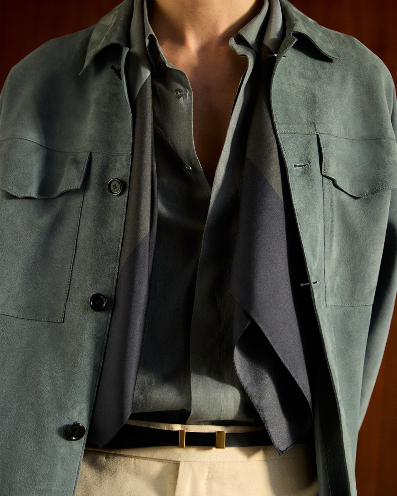 Close-up of torso wearing green suede jacket over an open shirt and navy scarf, beige trousers with black belt and gold buckle