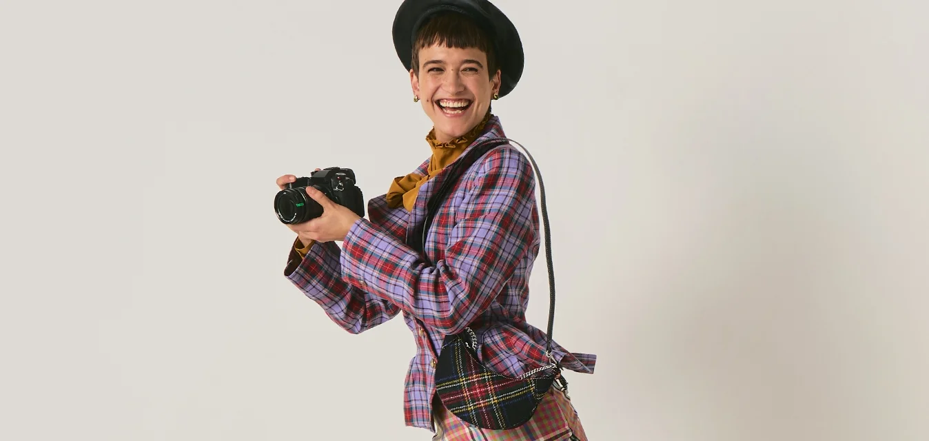 Giovane donna con outfit a quadri e stivali neri tiene una fotocamera, posando giocosa con una gamba sollevata contro uno sfondo uniforme.