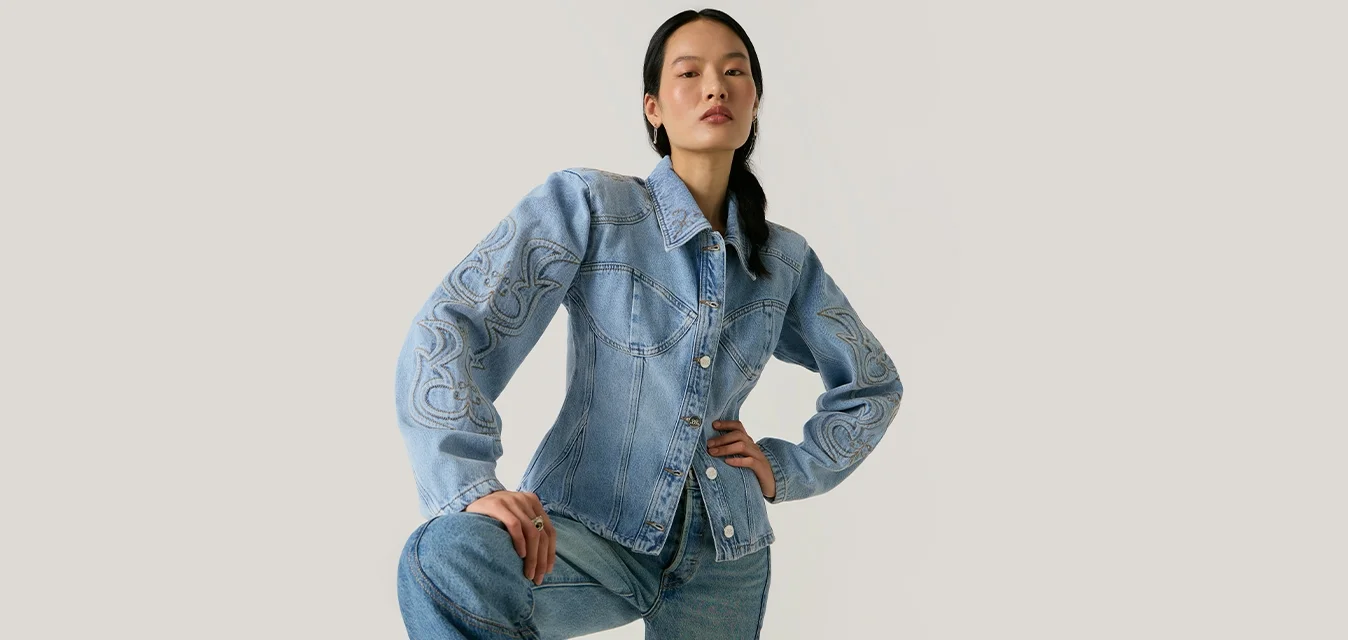 Donna con capelli lunghi e scuri che indossa una giacca di jeans azzurro chiaro ricamata e jeans abbinati, posando con un ginocchio sollevato davanti a uno sfondo semplice.