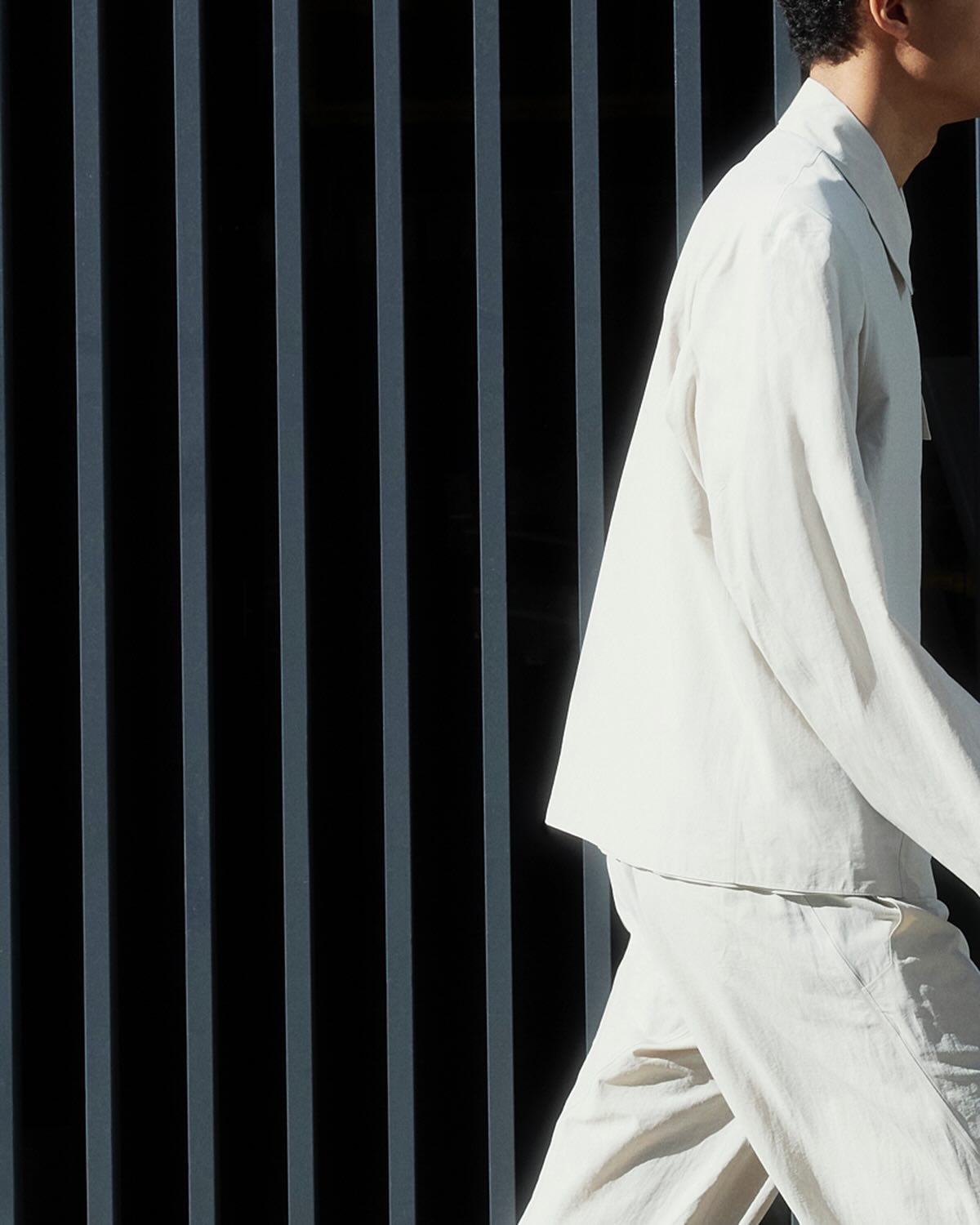 Partial view of person walking right in a white linen suit against vertical dark metal bars background