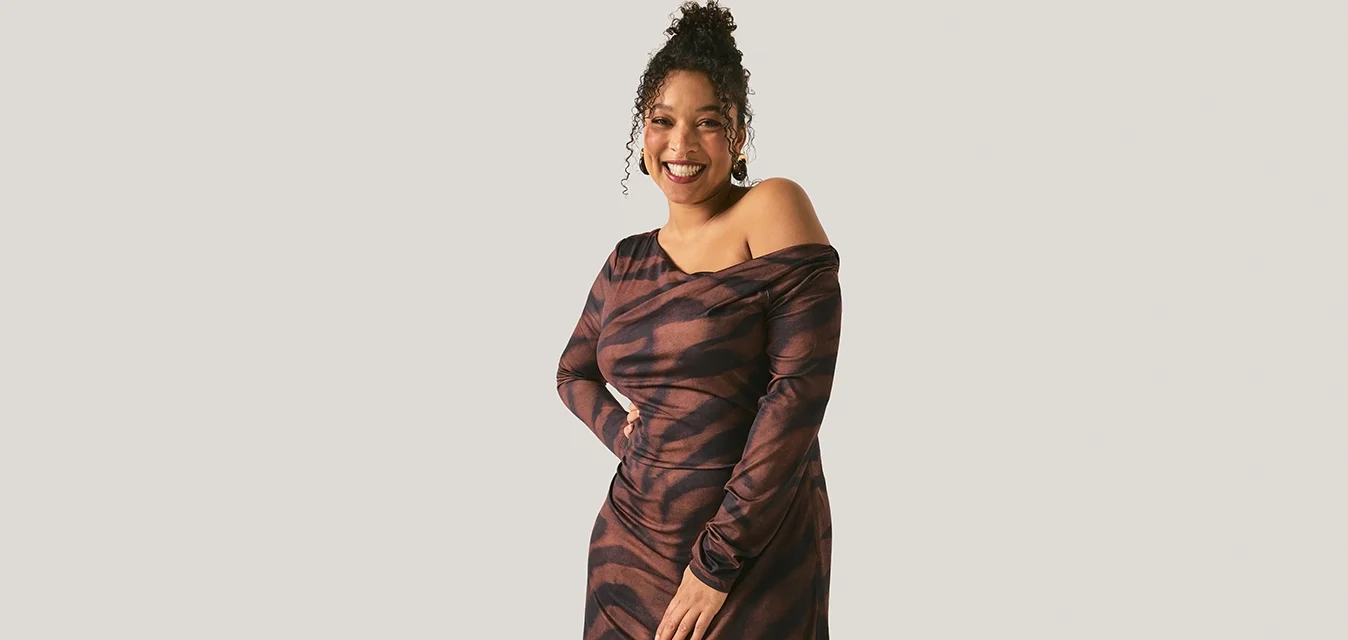 Donna sorridente con capelli ricci, vestita con un abito marrone e nero con motivo a fantasia e spalle scoperte, in piedi davanti a uno sfondo chiaro e uniforme.