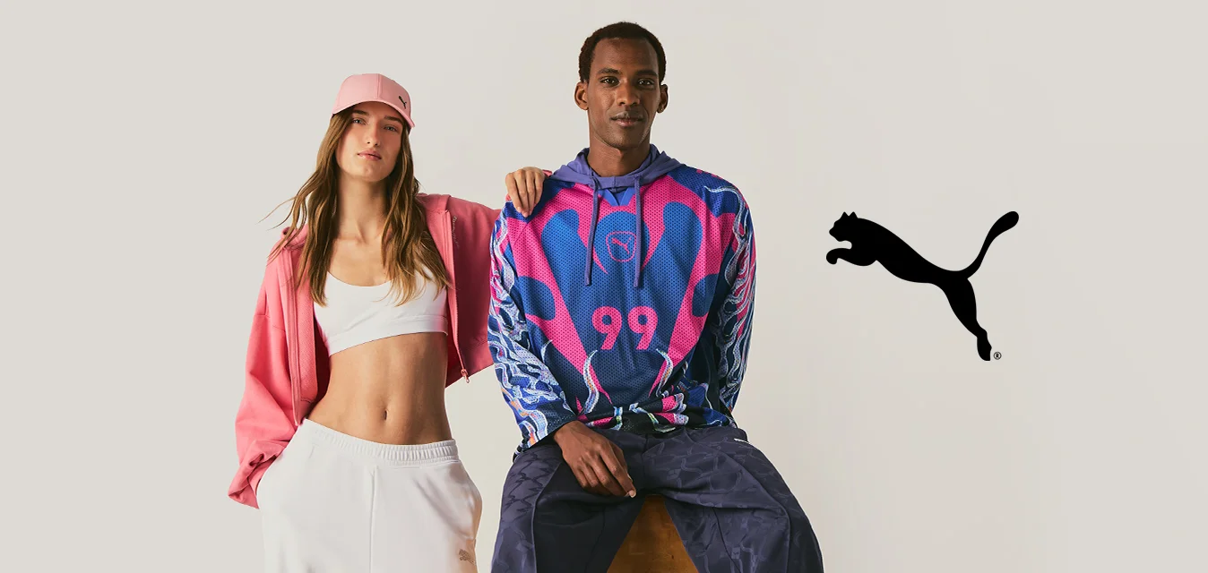 Una donna con pantaloni bianchi, crop top e giacca rosa sta accanto a un uomo seduto su uno sgabello che indossa una felpa con cappuccio colorata e pantaloni scuri.