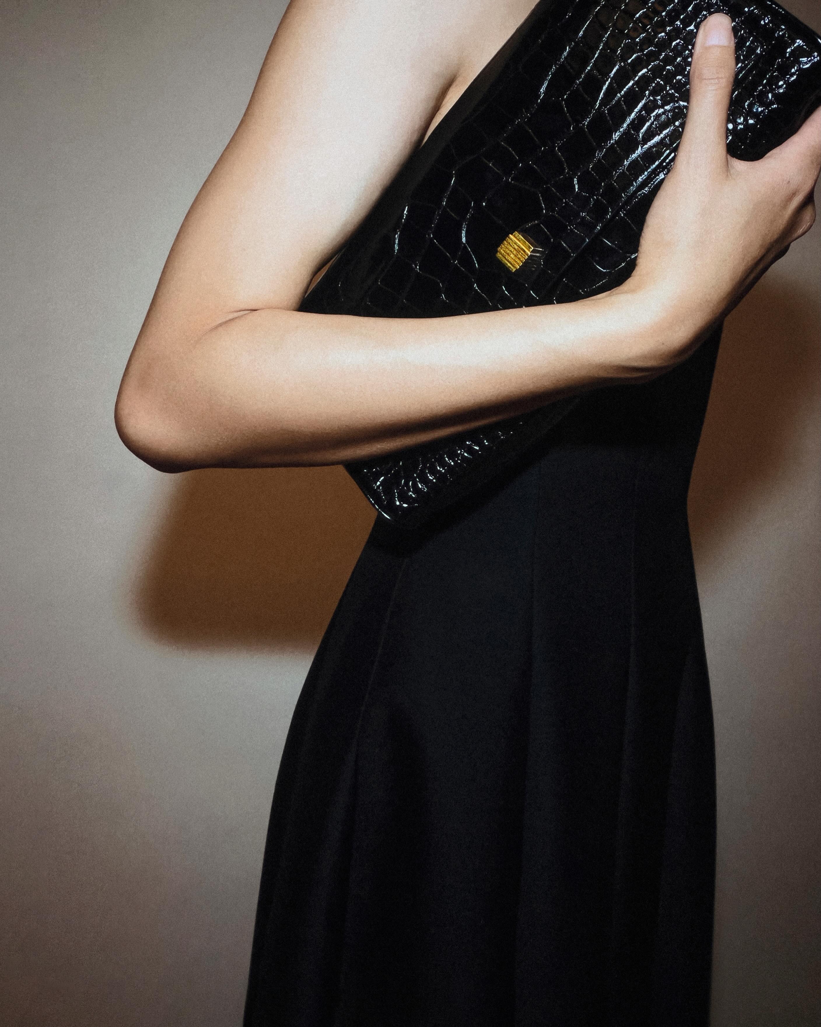 Cropped view of person in an elegant black dress holding a black crocodile‑pattern leather clutch bag with a gold clasp