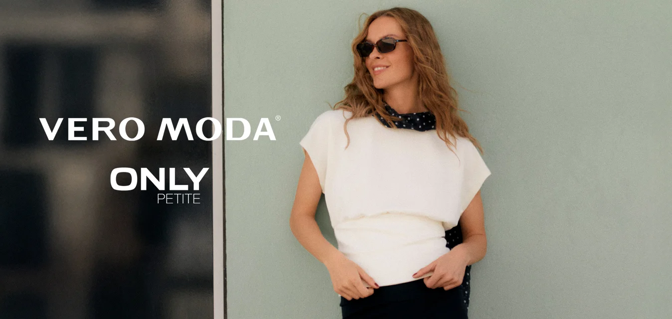 Donna con capelli mossi che indossa occhiali da sole, top bianco, pantaloni capri neri e una sciarpa nera con pois bianchi, che posa contro una parete verde chiaro.