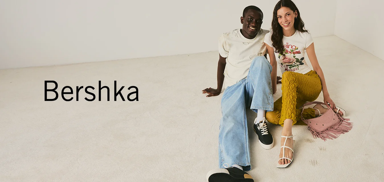 Due giovani seduti sul pavimento, uno indossa una camicia bianca e jeans azzurri, l’altro una t-shirt grafica e pantaloni color senape con una borsa rosa.