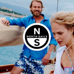 Due persone su una barca a vela vicino alla costa, una donna in bikini floreale e copricostume bianco che si muove con il vento, un uomo con camicia blu e pantaloncini a fantasia che sorride.