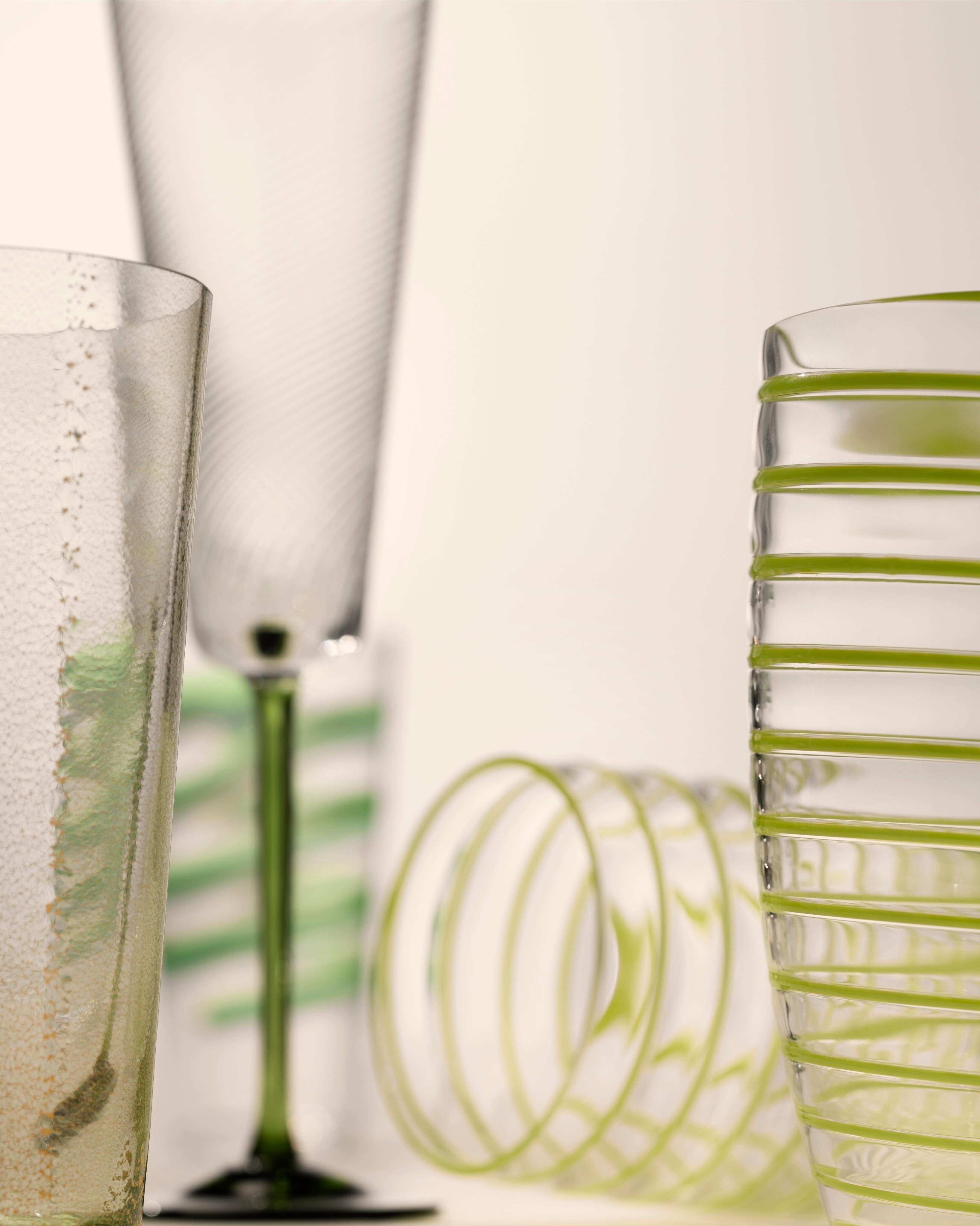 Close-up of decorative green-striped glassware: textured tumbler, tall champagne flute and stacked spiral-patterned glasses on neutral background