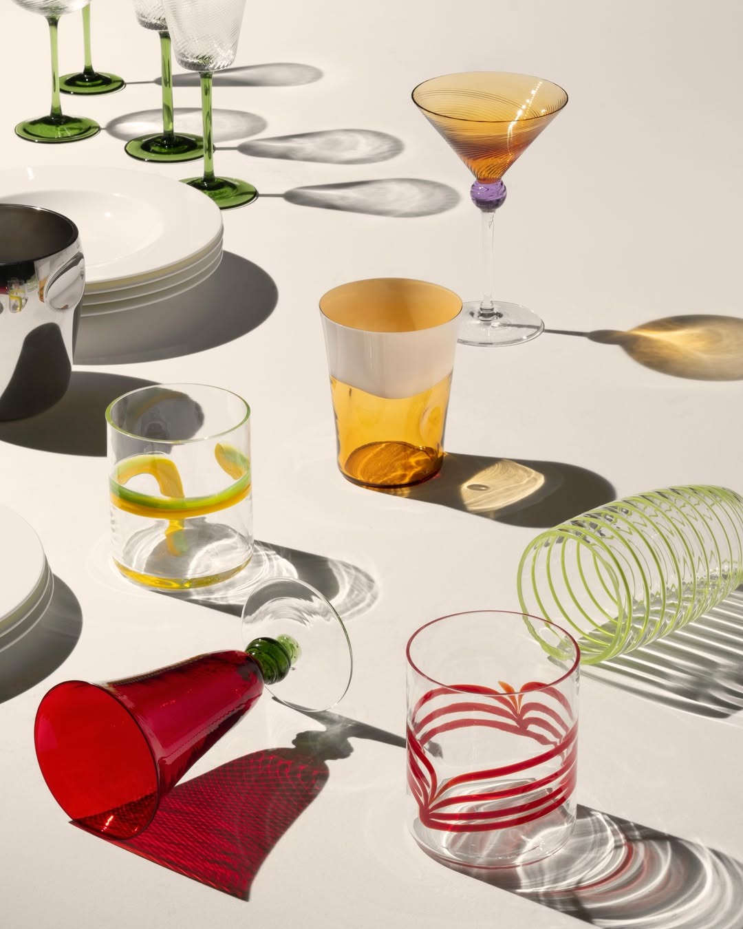 Colourful glassware — martini, tumblers and green‑stemmed wine glasses, a red goblet on its side casting shadows on white.