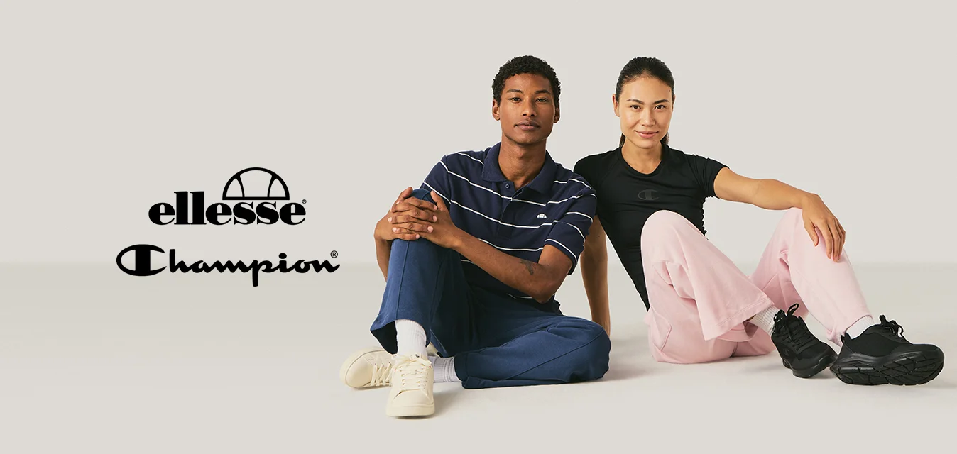 Due giovani adulti seduti per terra, uno con una camicia a righe e sneakers, l'altro con una camicia nera e pantaloni rosa.