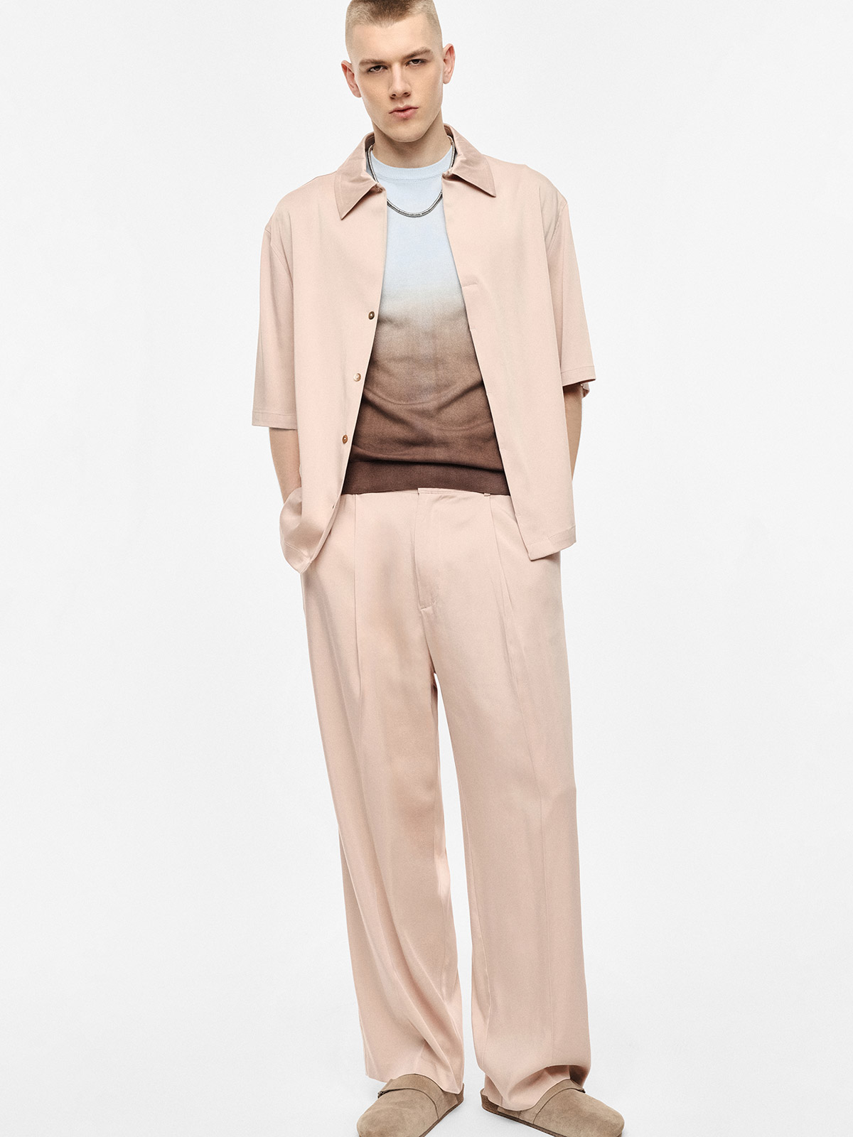 Male model wearing pastel pink short-sleeve shirt and wide trousers, ombre blue-brown knit, chain necklace and beige shoes on white background