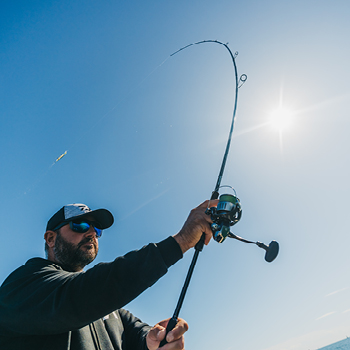 Fisherman wearing cap and sunglasses casting a fishing rod and reel against bright blue sky with sun