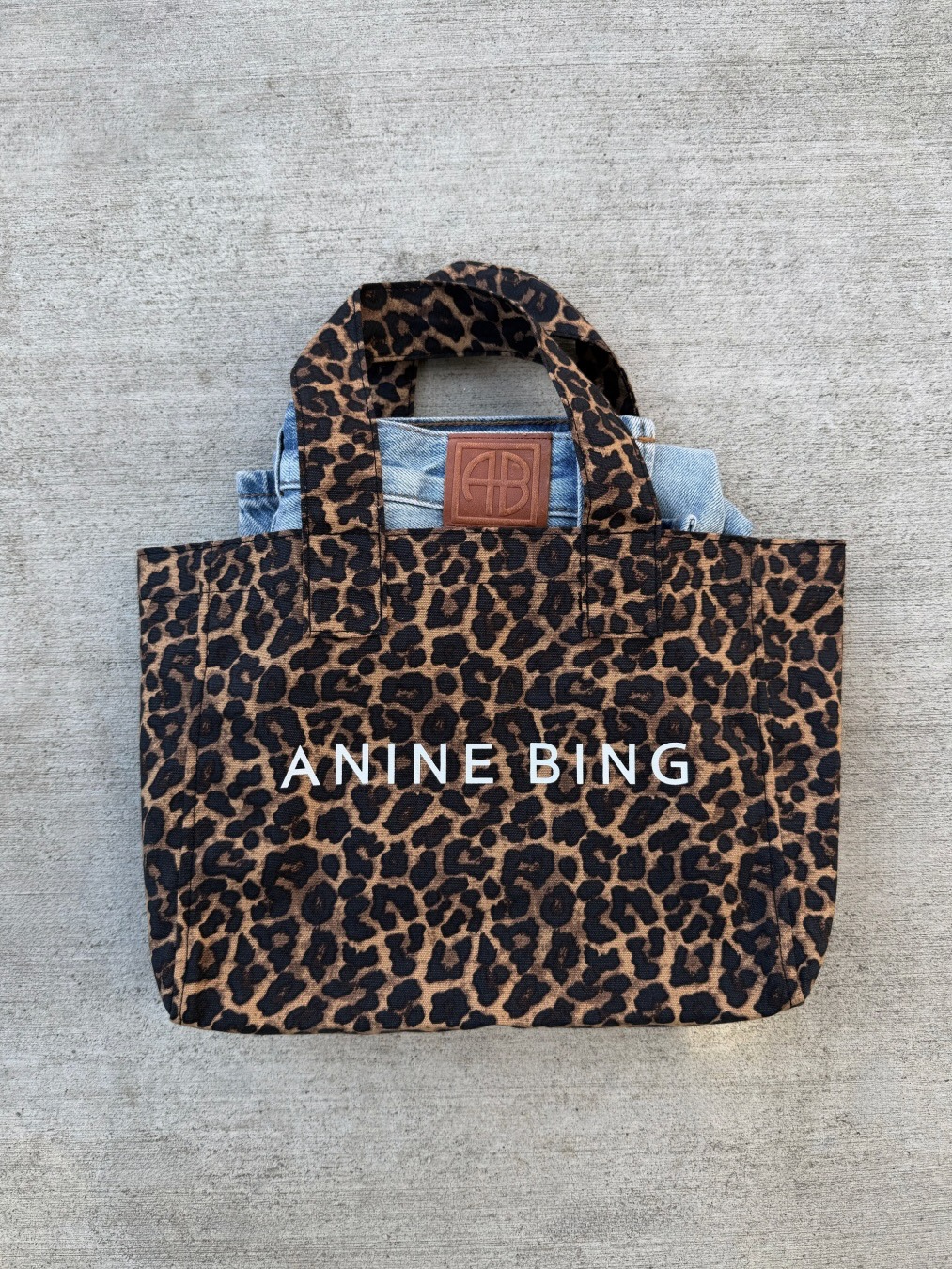Leopard-print Anine Bing canvas tote bag with white logo, denim jacket peeking out, photographed on concrete
