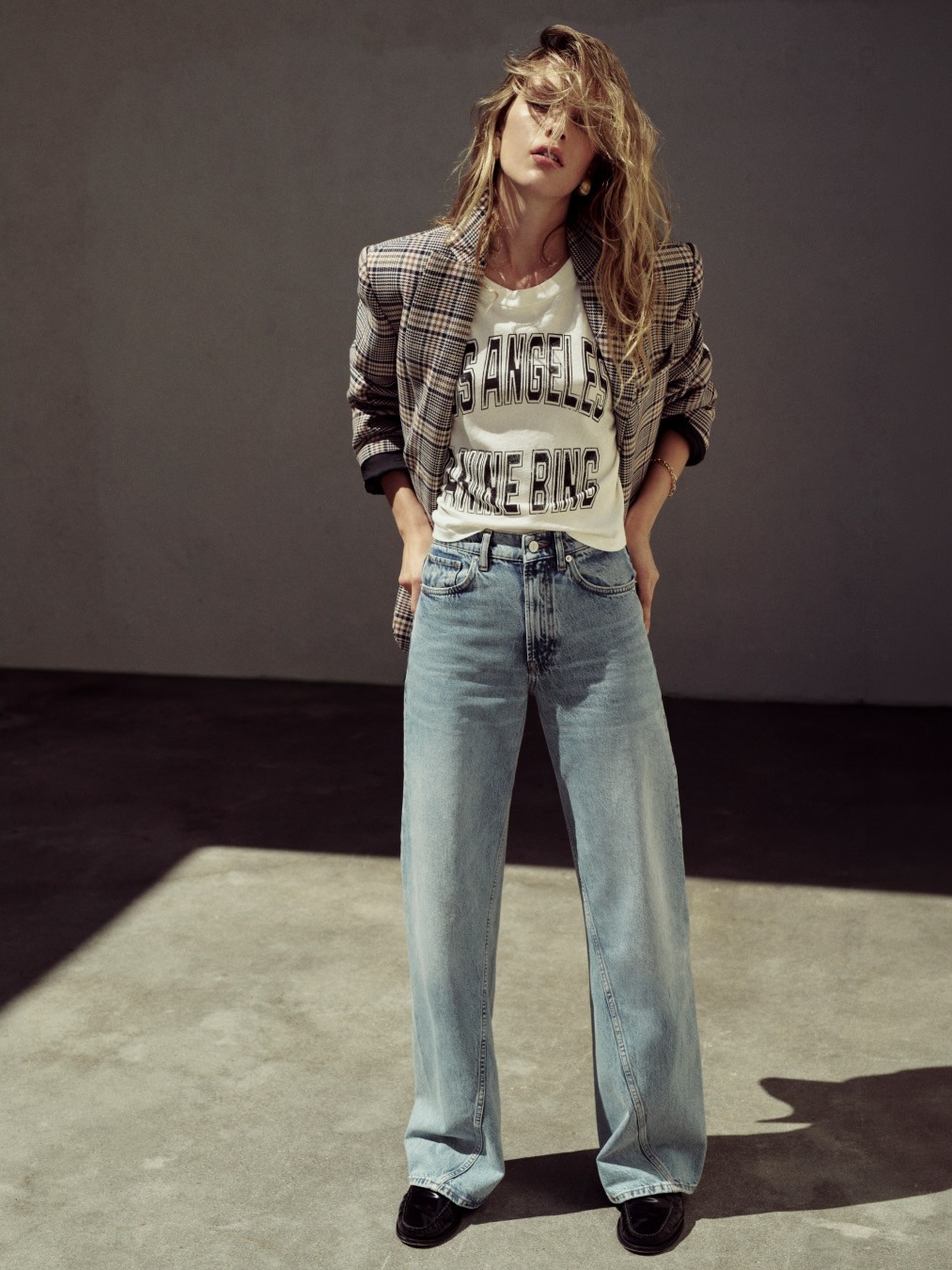Model wearing checked blazer, graphic 'Los Angeles' tee and high-waisted wide-leg jeans, hands in pockets in studio light