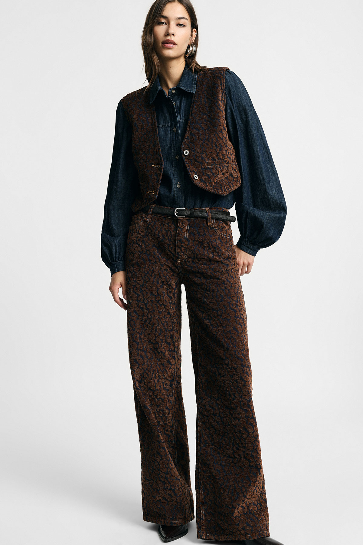 Woman model wearing brown patterned vest and matching wide-leg trousers with navy denim shirt and black belt, standing on white background