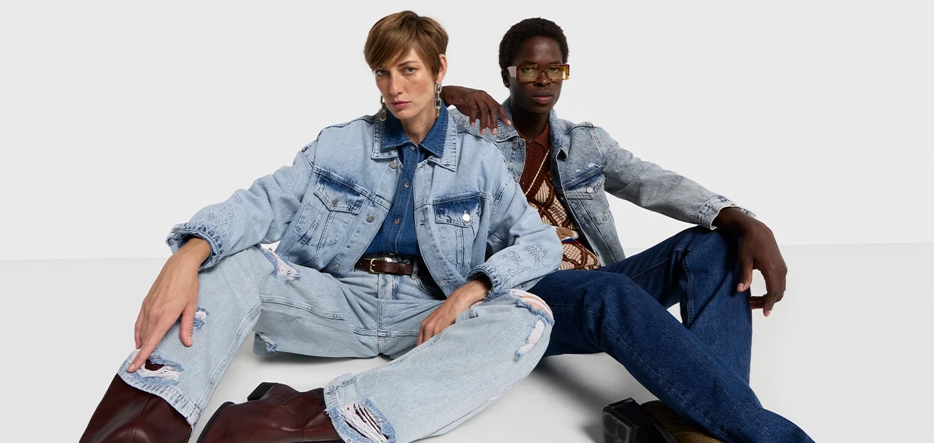 Due modelli seduti sul pavimento, indossano giacche di denim e jeans strappati. Il primo modello ha i capelli castani, il secondo indossa occhiali e una maglietta con motivo.
