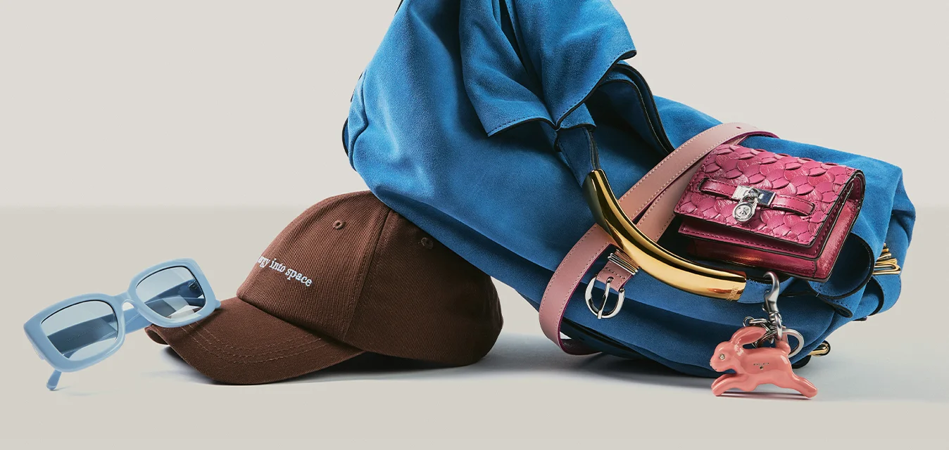 Borsa blu con portafoglio rosa e ciondolo sulla maniglia, accanto a un cappellino da baseball marrone e occhiali da sole blu su una superficie piana.