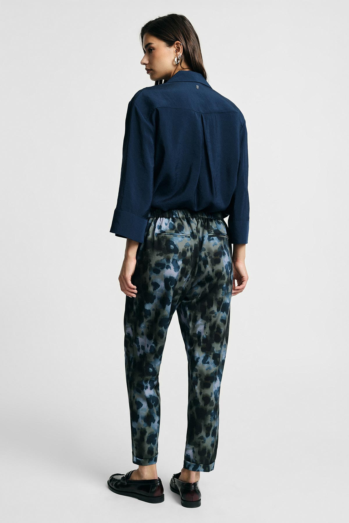 Young woman seen from behind wearing a navy blouse, elastic-waist blue-green patterned trousers and black loafers on a grey background