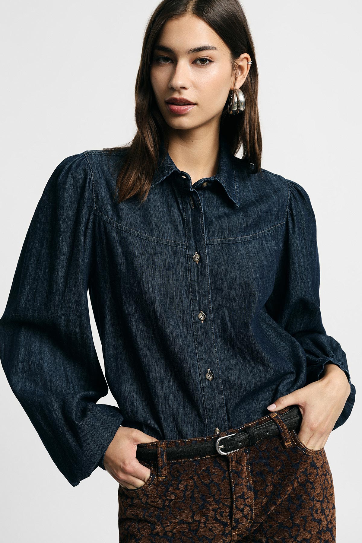Woman wearing dark denim button-up blouse with puffed sleeves and patterned brown trousers, hands in pockets