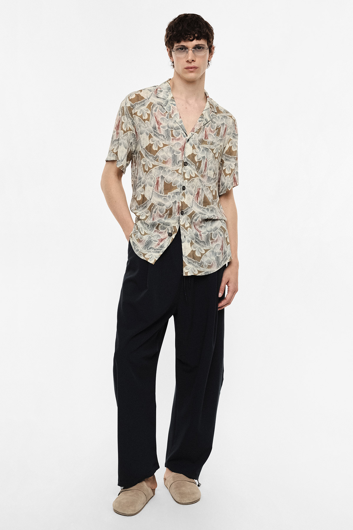 Male model wearing patterned short-sleeve button shirt, black wide-leg trousers and beige slip-on shoes, standing against white background