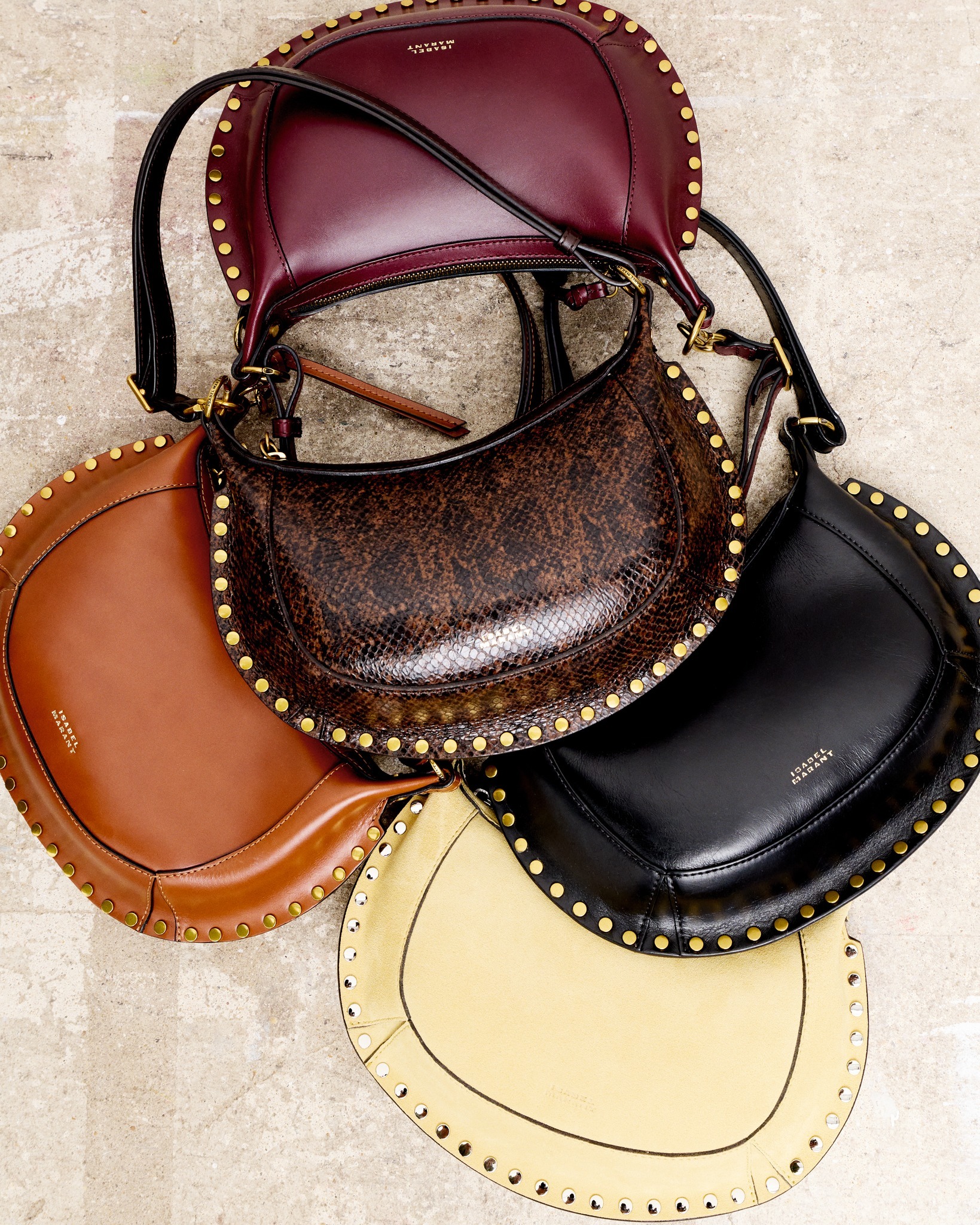 Five studded leather saddle bags in burgundy, brown, black, tan and cream arranged on a textured stone surface