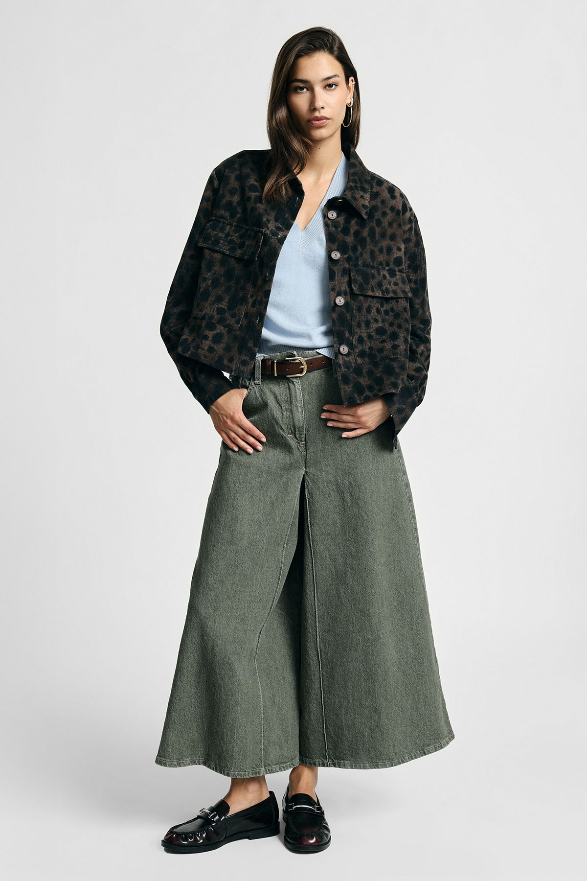 Woman wearing leopard-print cropped jacket, pale blue V-neck top, wide-leg green denim culottes, brown belt and black loafers on plain background