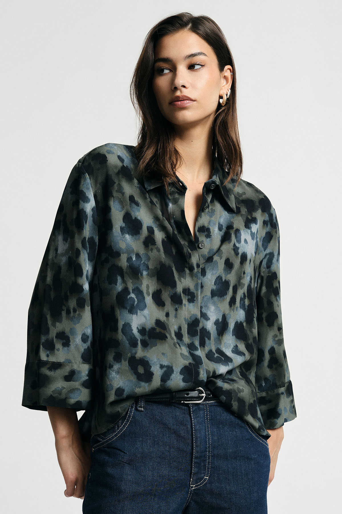 Woman in green-black leopard-print satin blouse tucked into dark blue jeans against plain light background