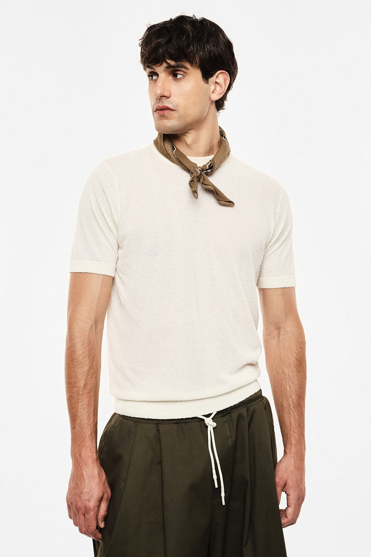 Male model wearing cream short-sleeve knit top, olive green drawstring trousers and khaki neckerchief, standing against white background