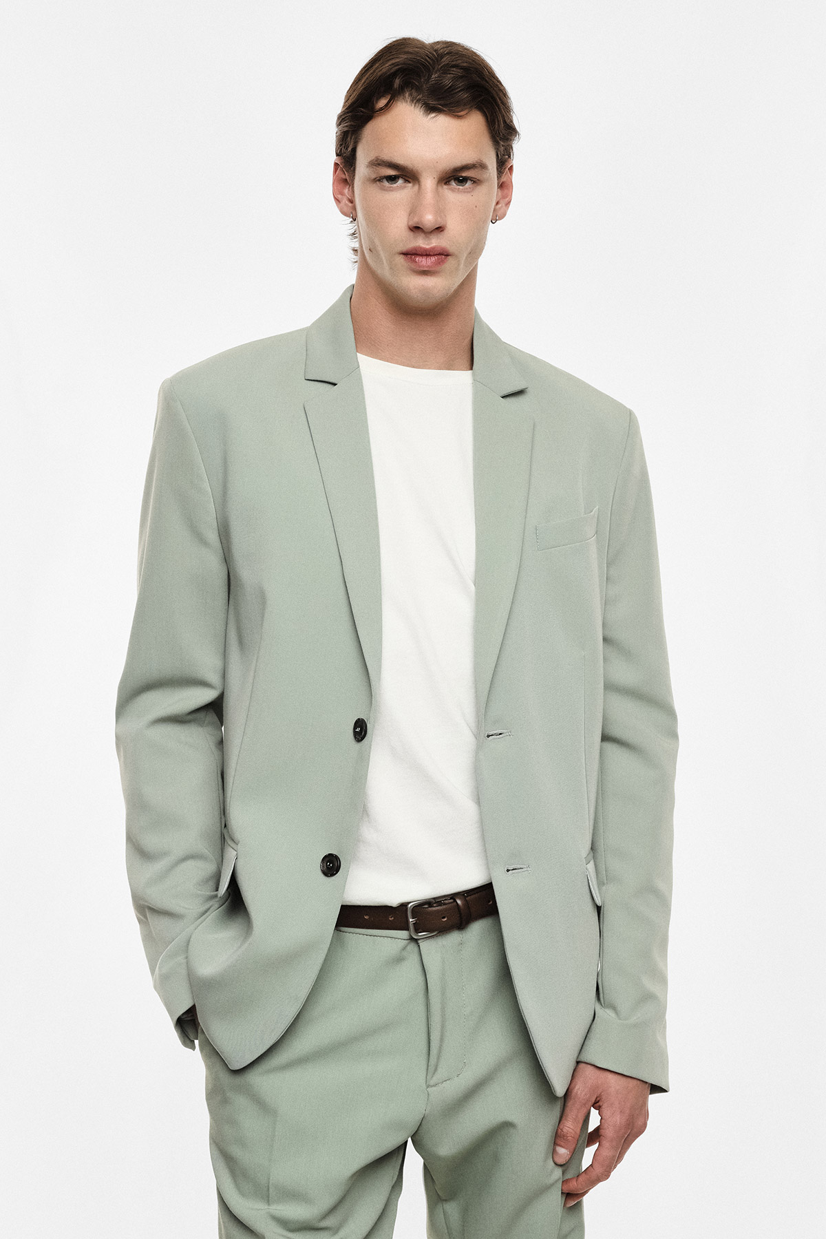 Male model wearing a light sage suit with white T-shirt and brown belt, standing against a plain white background