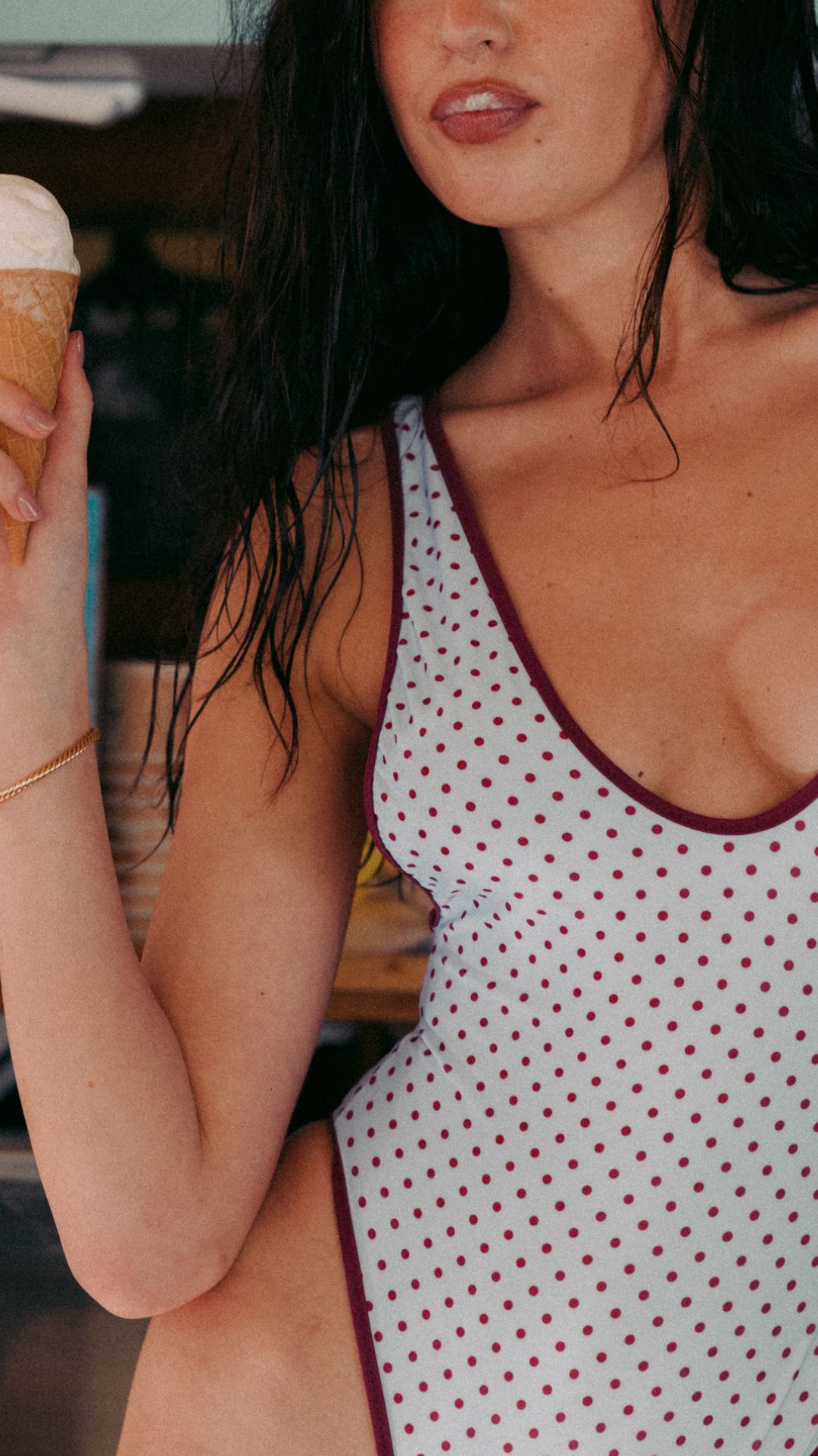 Close-up of a woman in a white polka-dot swimsuit holding an ice cream cone, cropped at chin and torso, summery scene