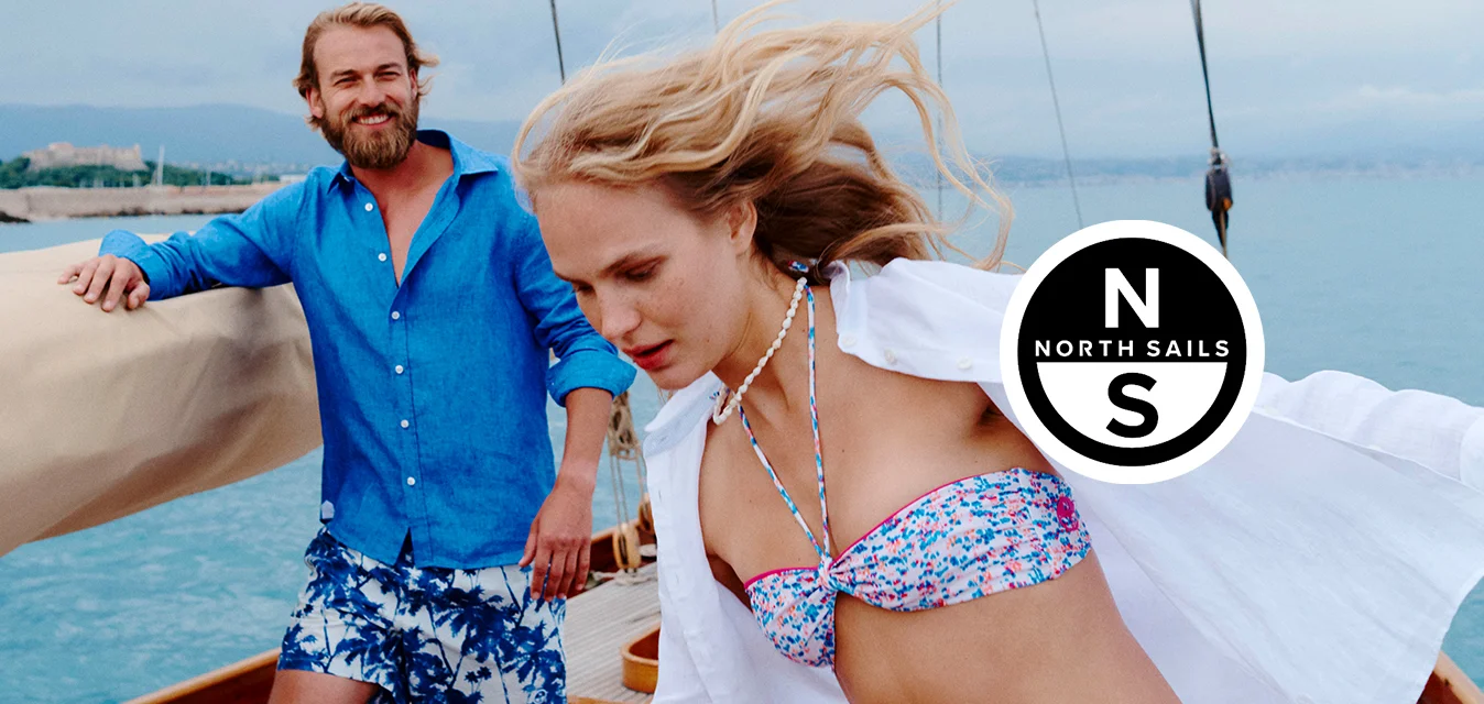 Due persone su una barca a vela vicino alla costa, una donna in bikini floreale e copricostume bianco che si muove con il vento, un uomo con camicia blu e pantaloncini a fantasia che sorride.