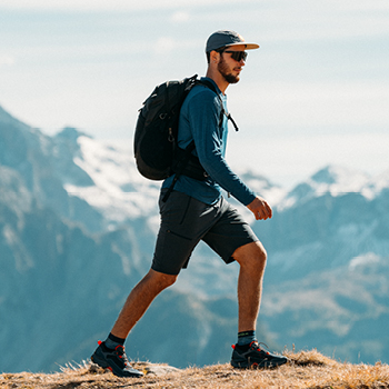 Man hiking with backpack and cap on alpine ridge, trekking boots and sunglasses, mountains in background