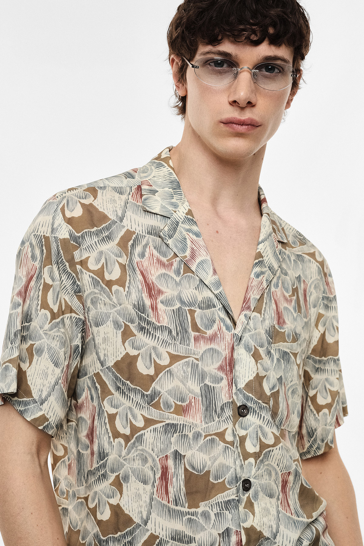 Young man wearing a patterned short-sleeve shirt and thin-rimmed glasses, waist-up on a white background