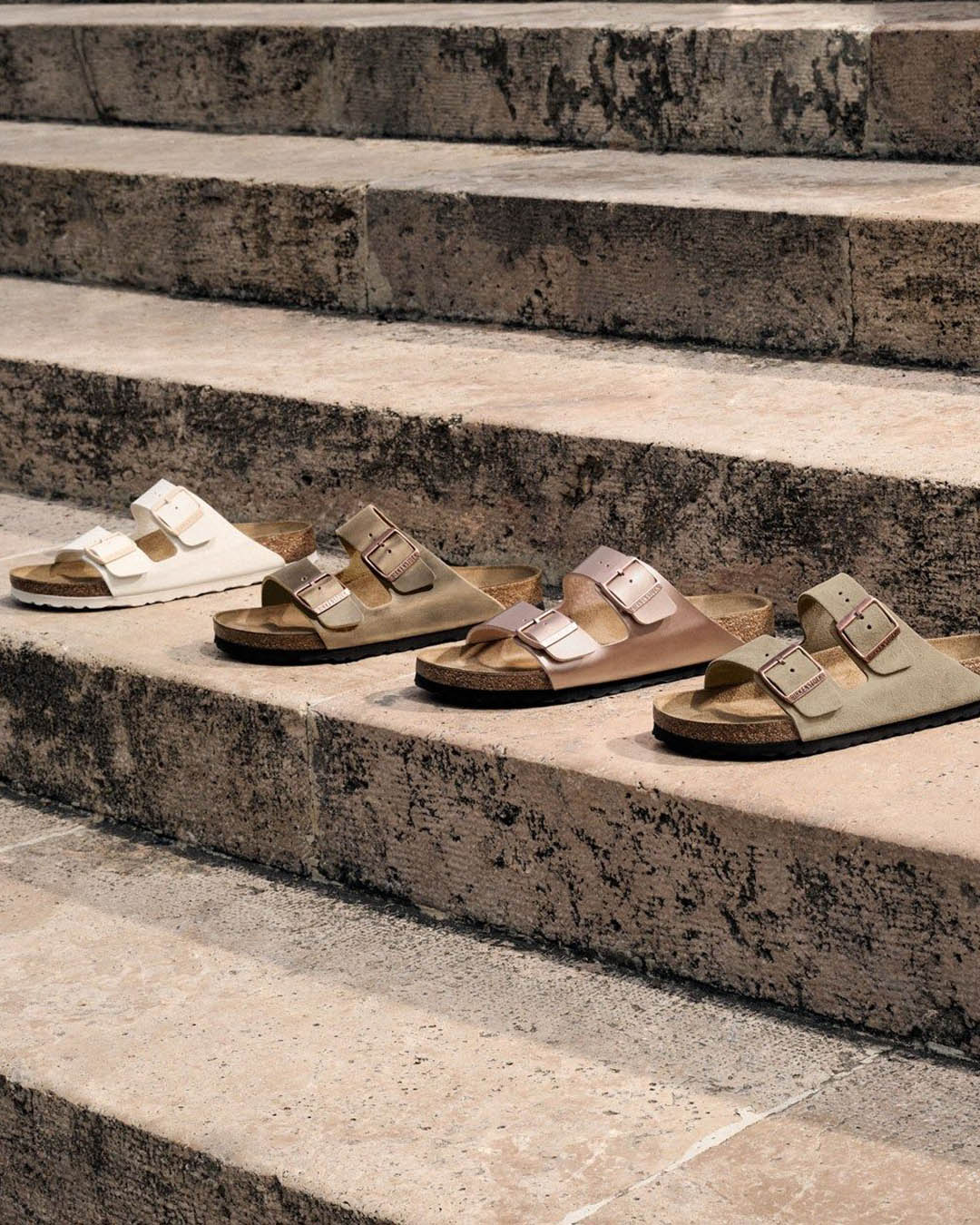 Four pairs of double-buckle sandals in neutral tones (cream, olive, tan, khaki) arranged on weathered stone steps