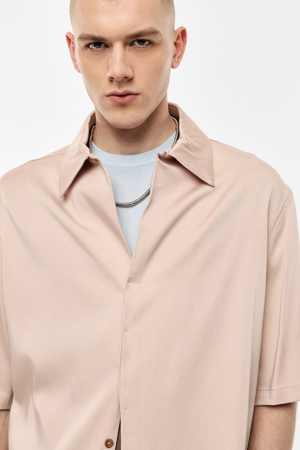 Close-up fashion portrait of a young man wearing a pale pink short-sleeve shirt and silver chain necklace against a plain white background