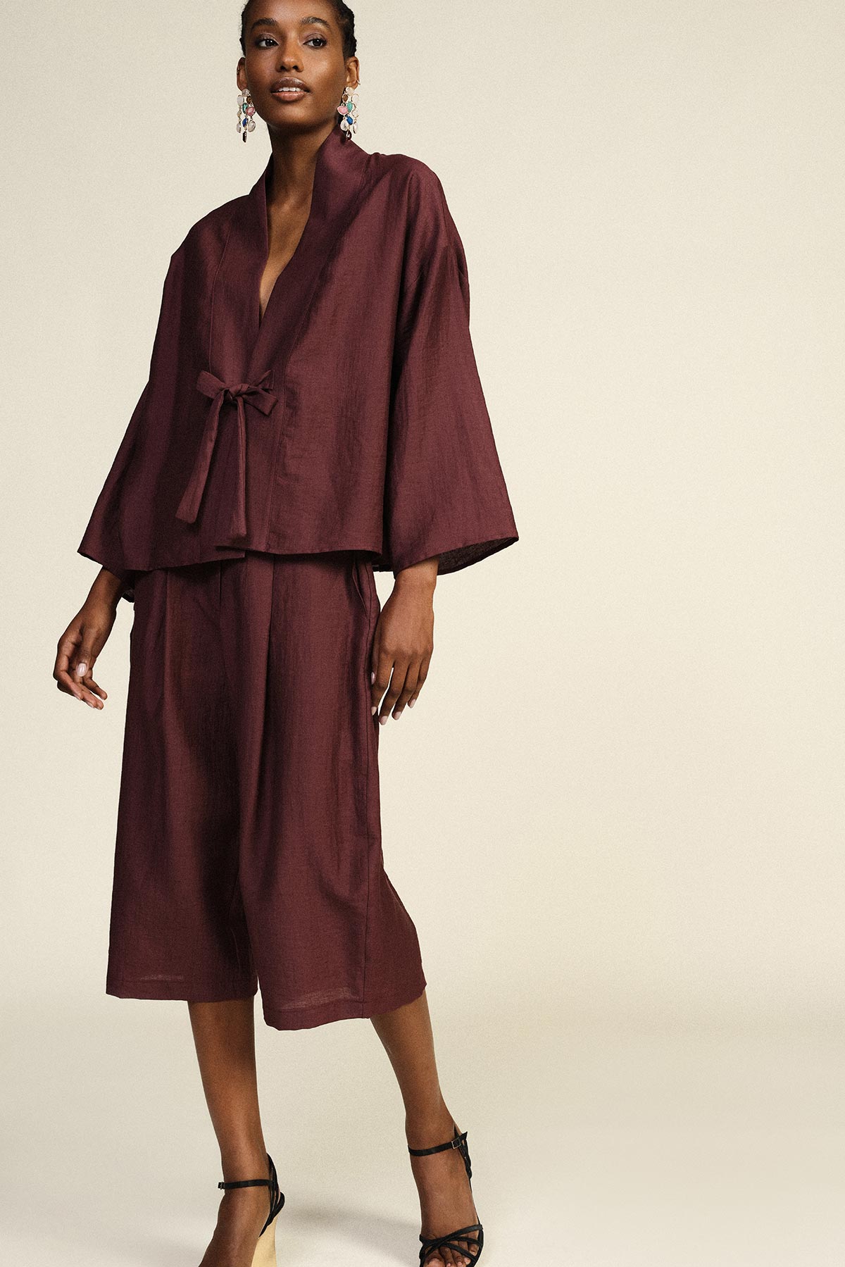 Woman modelling burgundy kimono-style jacket with tie and matching wide-leg cropped trousers and black strappy heels on neutral background