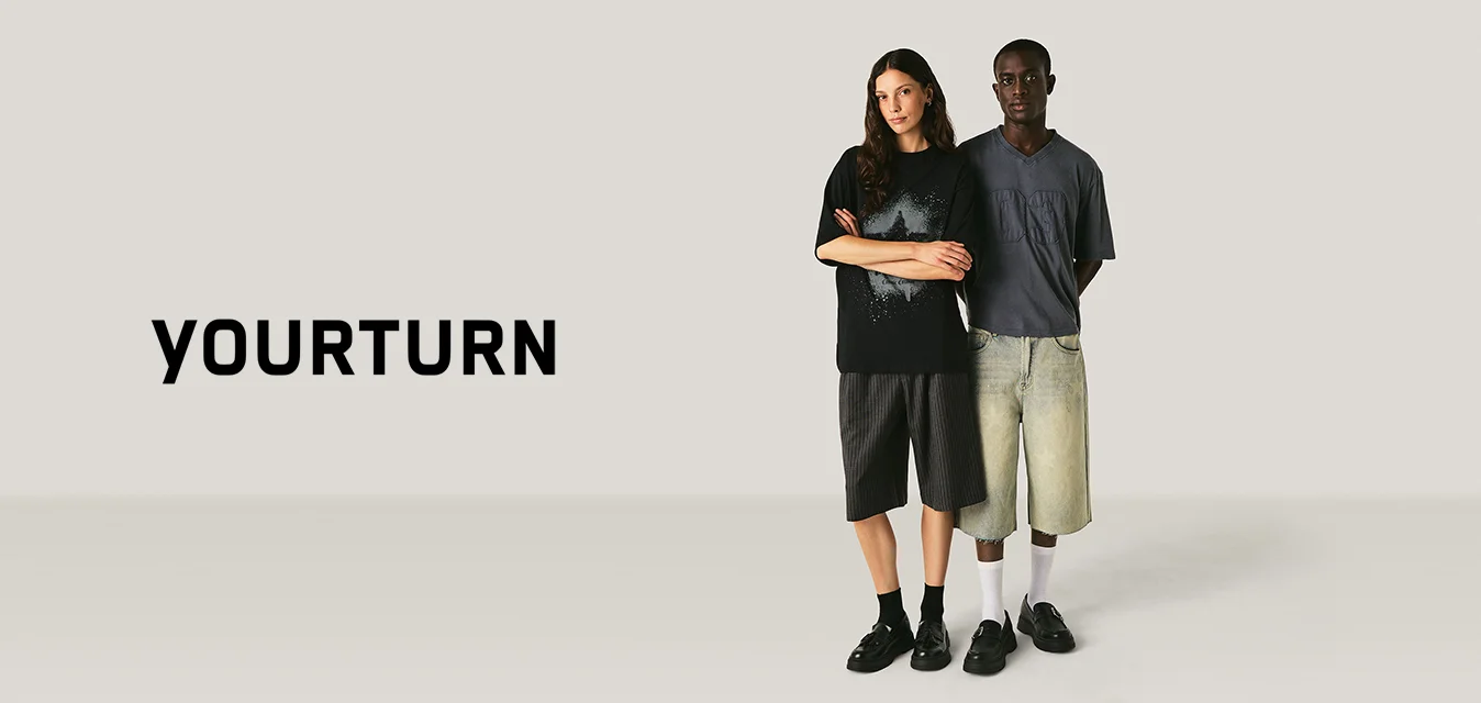 Due persone in piedi affiancate che indossano magliette oversize e pantaloncini al ginocchio su uno sfondo chiaro e uniforme.