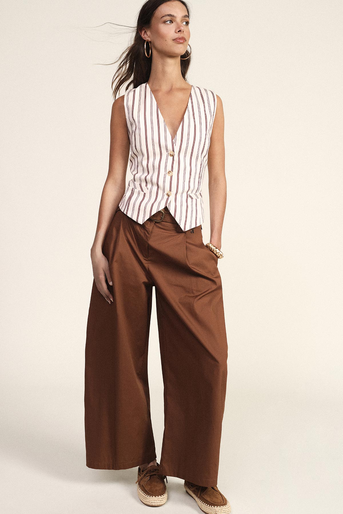 Young woman modelling sleeveless striped waistcoat, wide-leg brown trousers and espadrilles, gold hoop earrings and bracelet