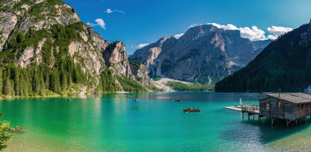 Turquoise alpine lake with wooden boathouse and rowboats, surrounded by forested mountains under a clear blue sky